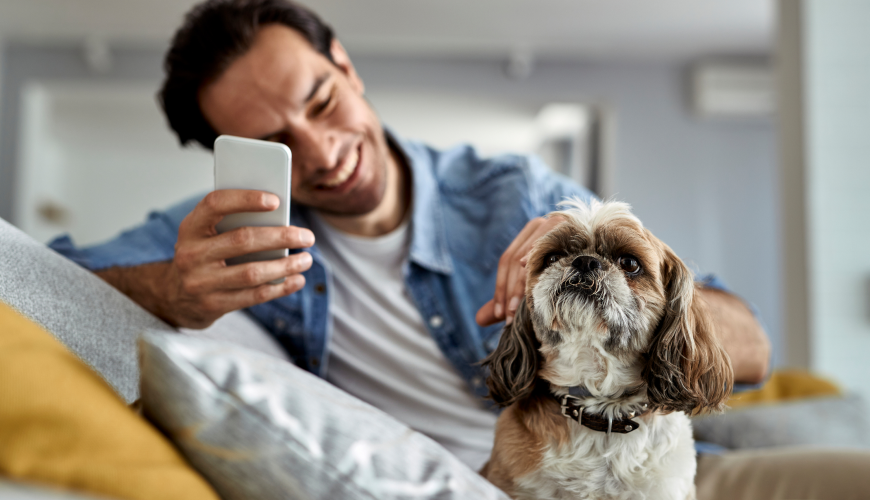 Smiling man on couch petting a small dog while holding his phone