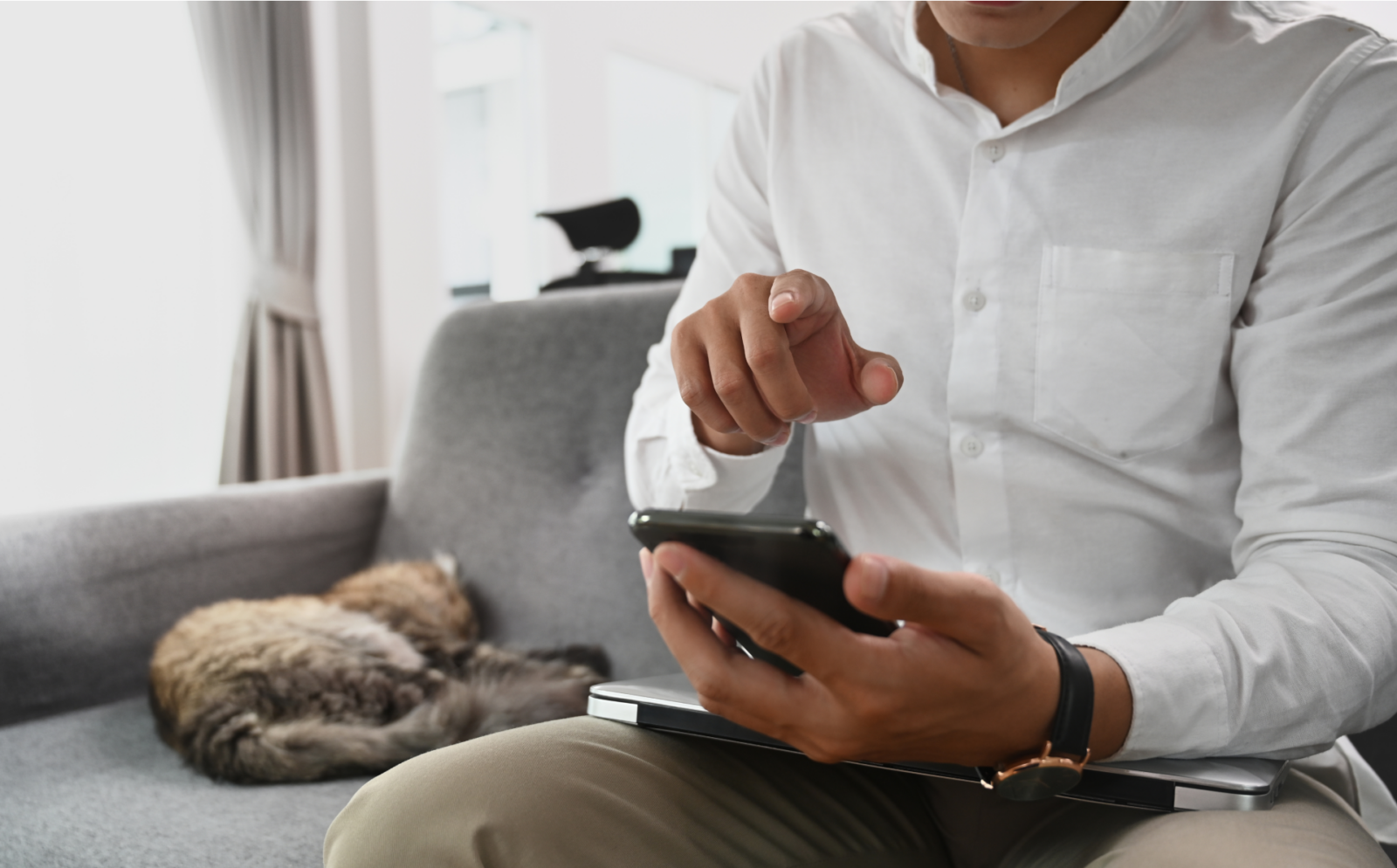 Man in white shirt using his mobile phone sitting on a couch with a cat sleeping nearby