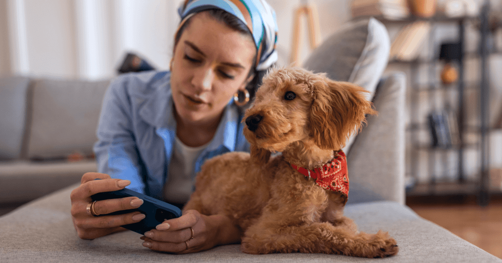 A young woman lying on her stomach using her mobile phone, with her dog relaxing next to her.