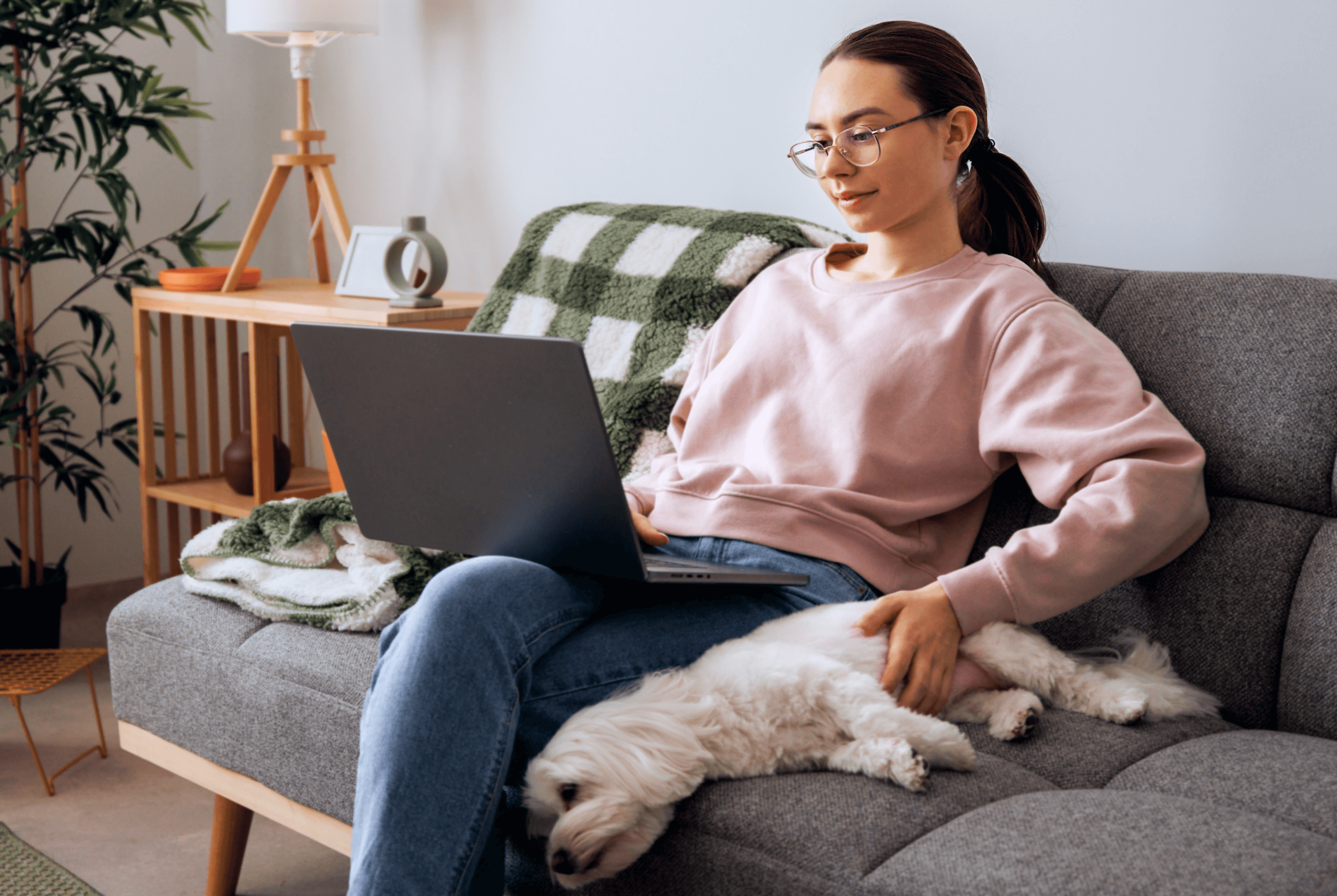 A woman using her laptop on her couch, petting her sleeping dog next to her.