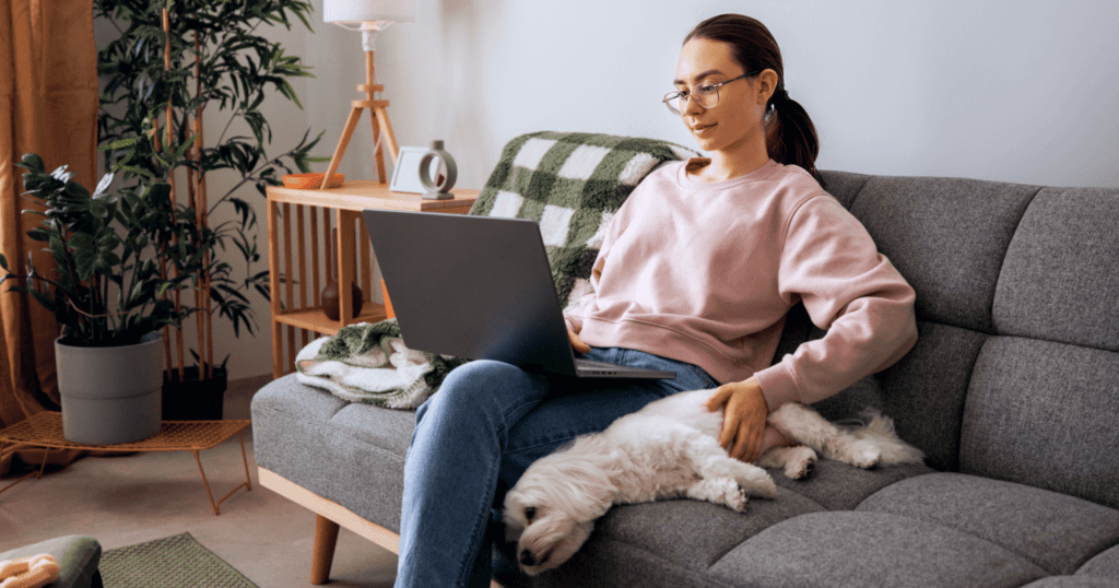 A woman using her laptop on her couch, petting her sleeping dog next to her.