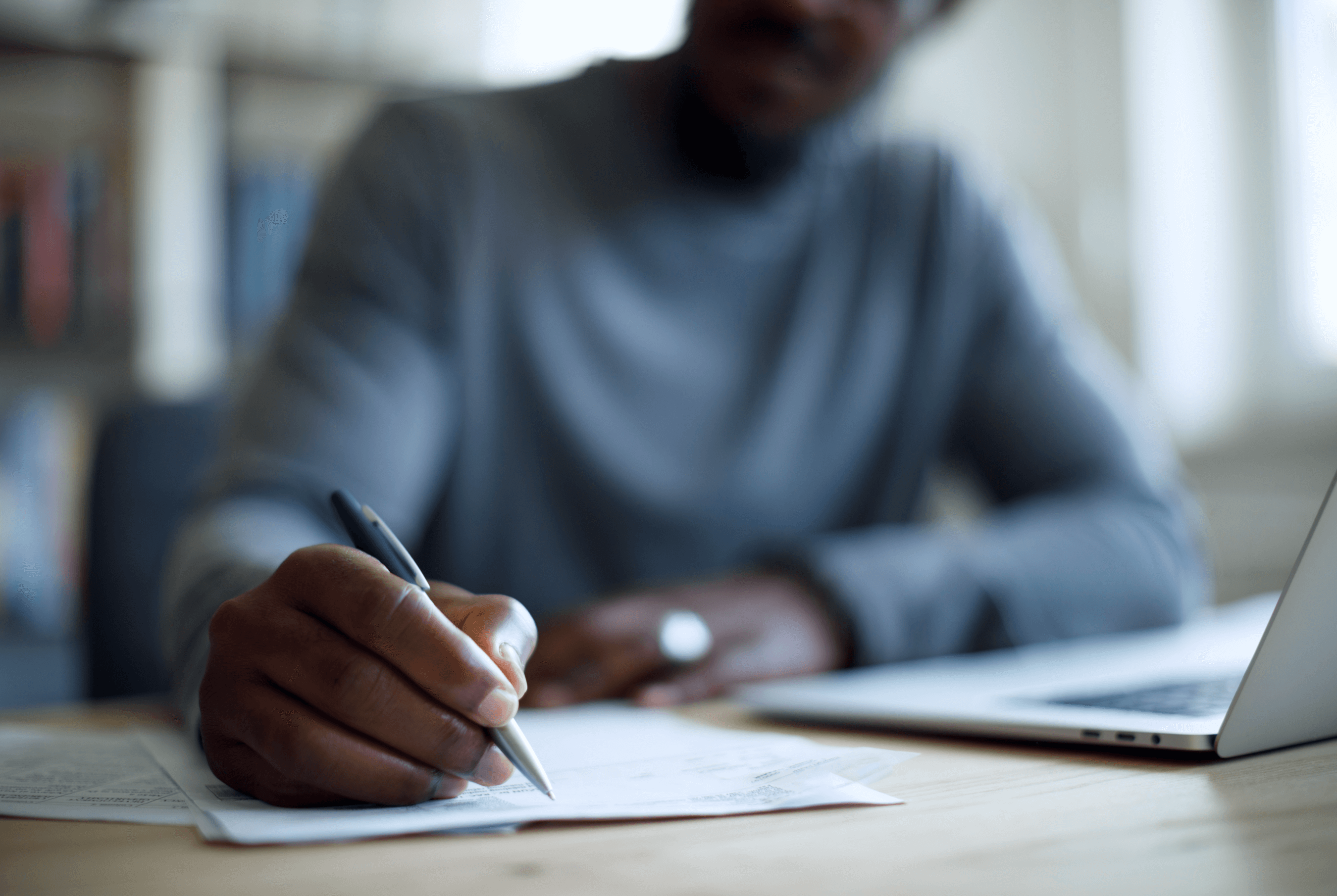 Closeup of a person writing on paper next to an open laptop, with their body and face blurred in the background