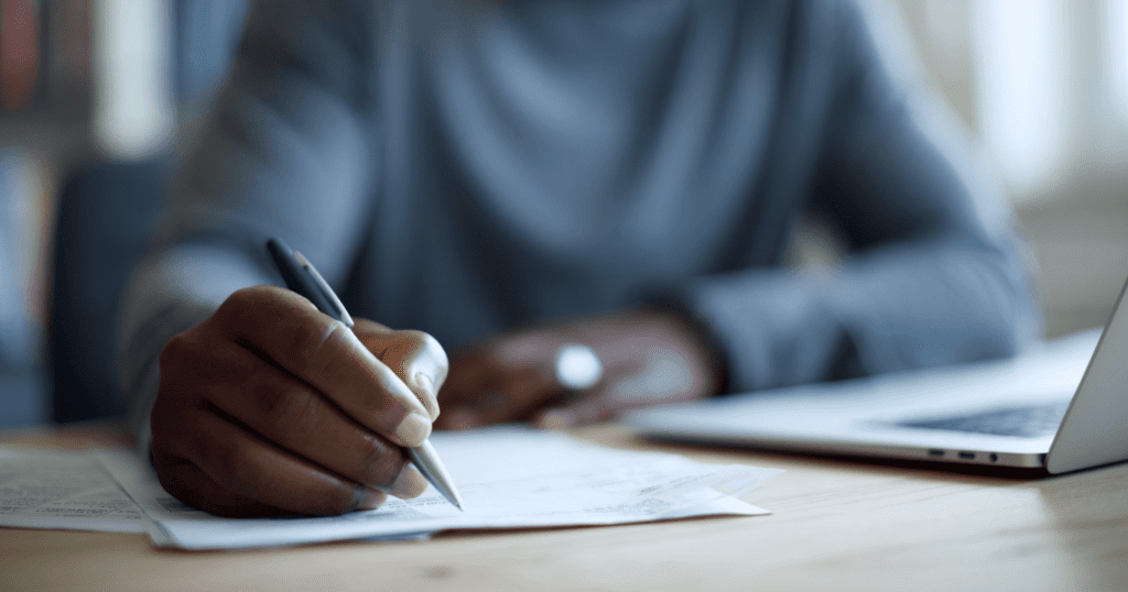 Closeup of a person writing on paper next to an open laptop, with their body and face blurred in the background