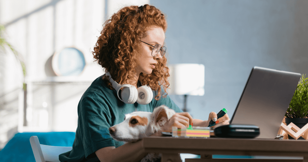 Young woman working on her laptop while her dog rests its chin on her arm.