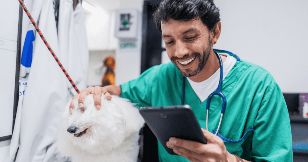 A young male vet tech petting a small white dog while smiling and reading a mini tablet.