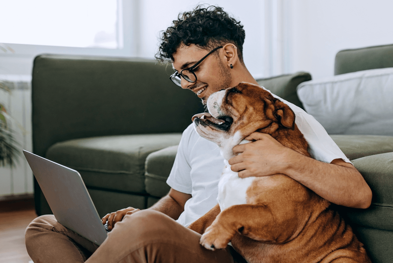 A smiling young man hugging his bulldog while using his laptop.