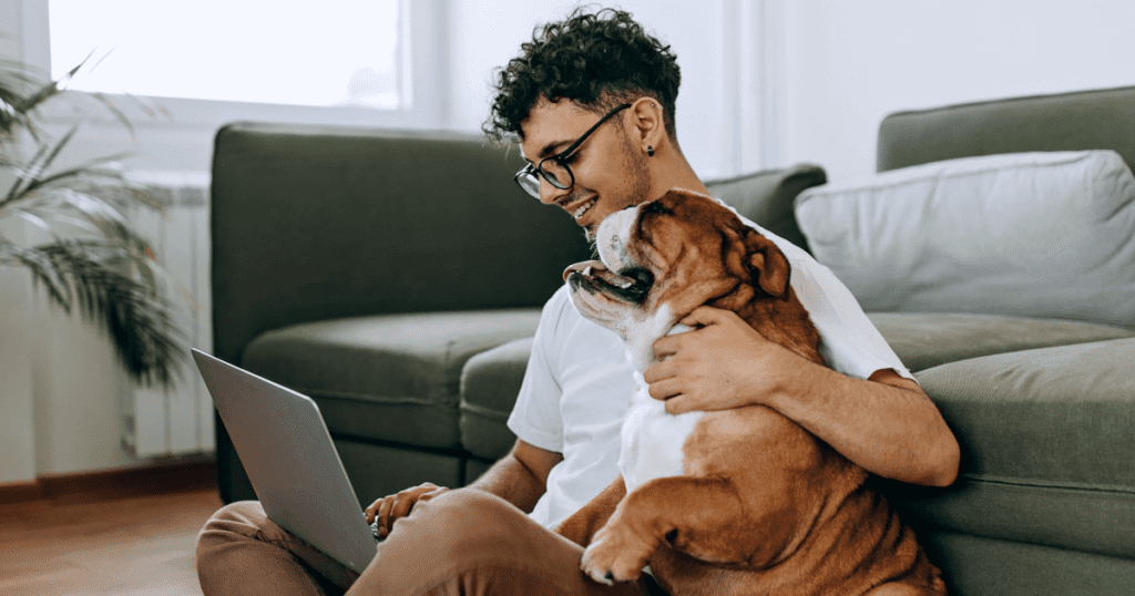 A smiling young man hugging his bulldog while using his laptop.