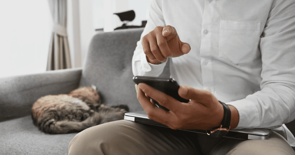 Man sitting on couch with closed laptop using his smartphone with cat curled up asleep beside him