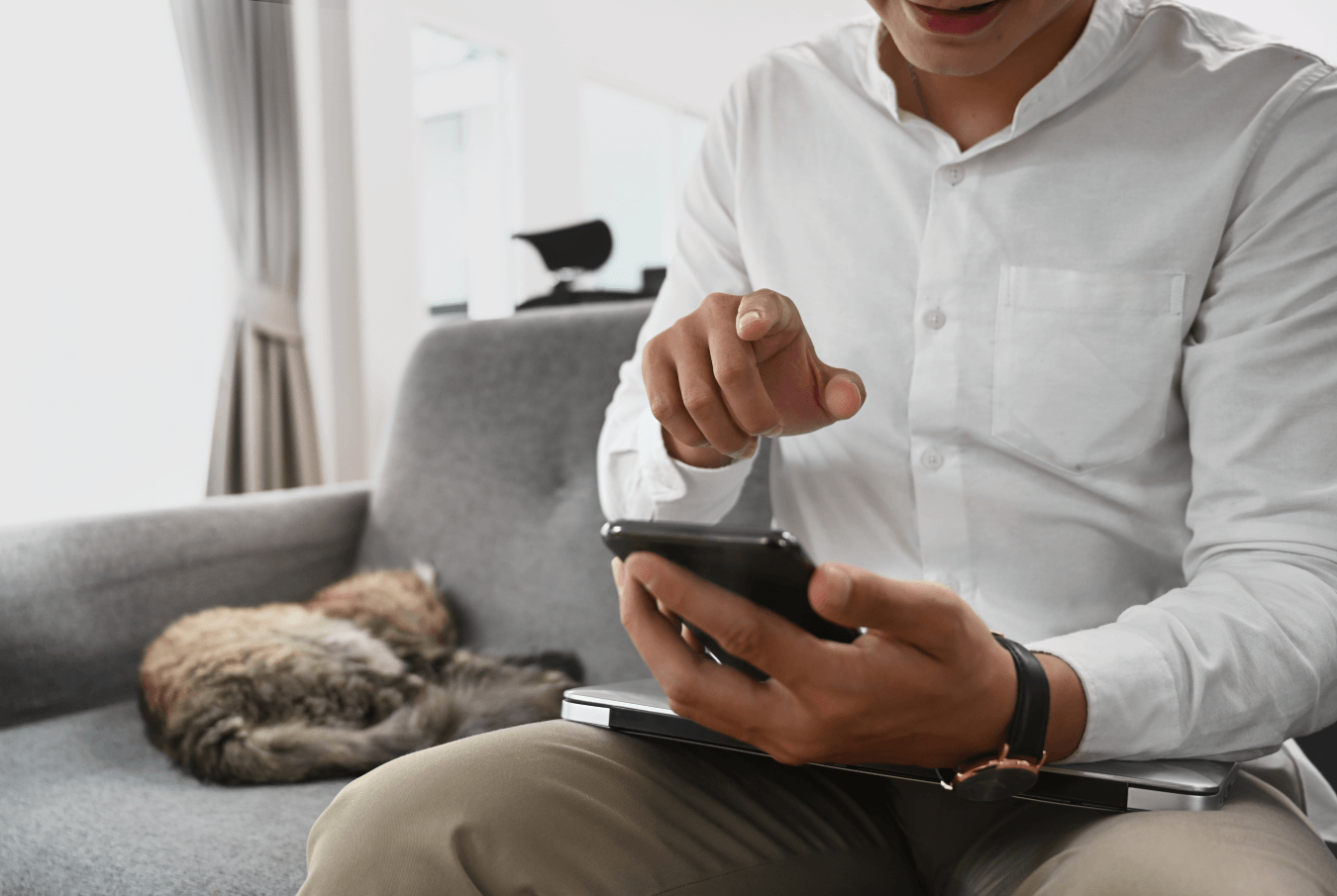 Man sitting on couch with closed laptop using his smartphone with cat curled up asleep beside him