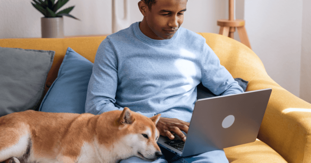 Smiling man using his laptop while sitting on the couch; his shiba inu rests its head on his lap