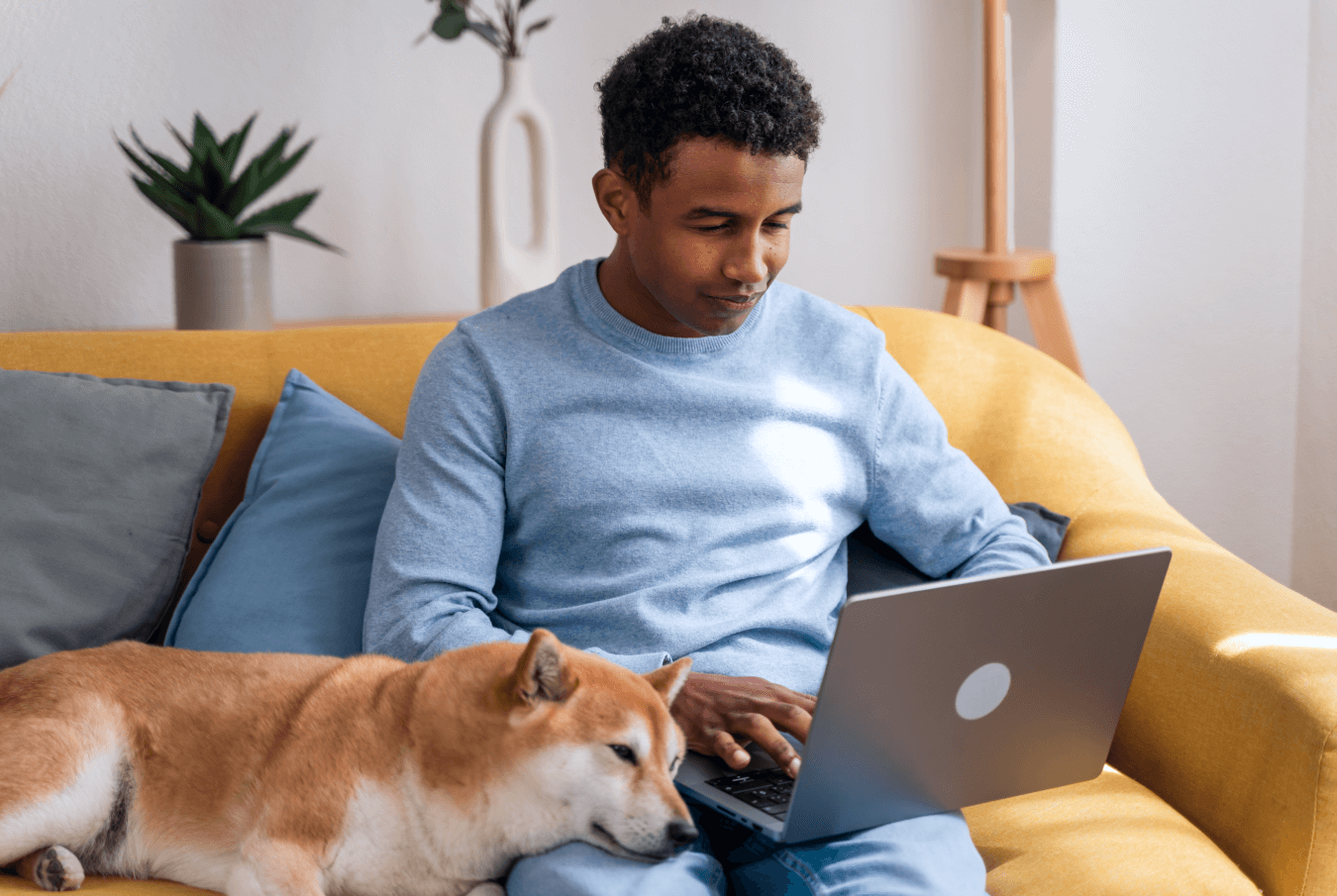 Smiling man using his laptop while sitting on the couch; his shiba inu rests its head on his lap