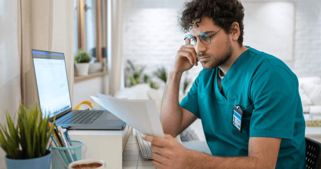 Thoughtful male vet looking at paperwork with his laptop open at his desk