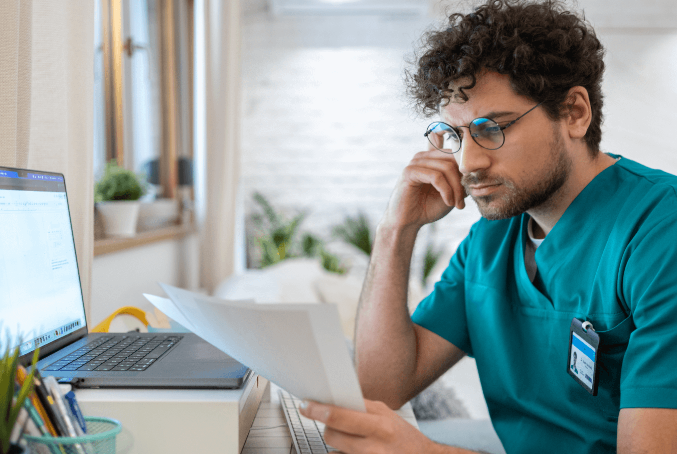 Thoughtful male vet looking at paperwork with his laptop open at his desk