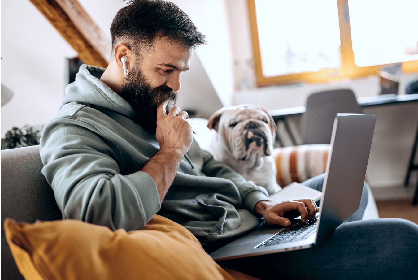 Smiling bearded pet owner uses his laptop with his bulldog sitting next to him on the couch