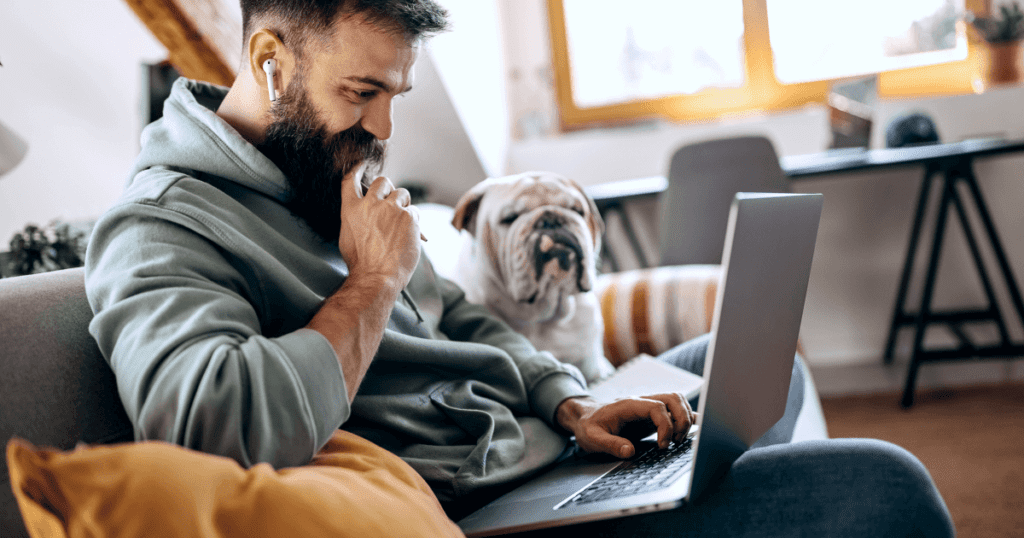 Smiling bearded pet owner uses his laptop with his bulldog sitting next to him on the couch