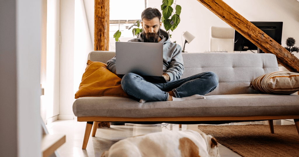 Pet owner sitting cross-legged on a couch with a bulldog laying on the floor at his feet.