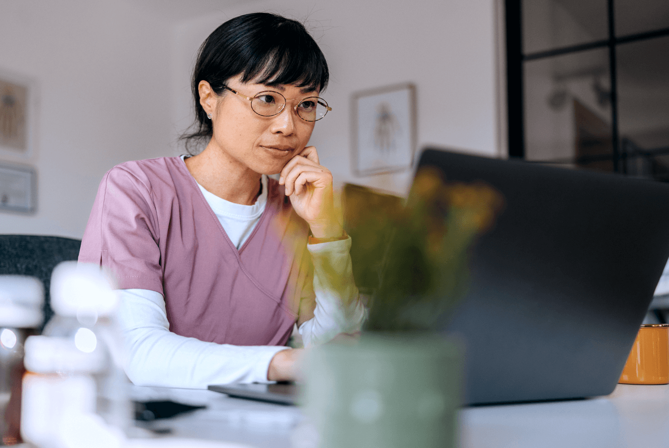 Woman in glasses and pink scrubs thoughtfully reviewing data on a laptop screen