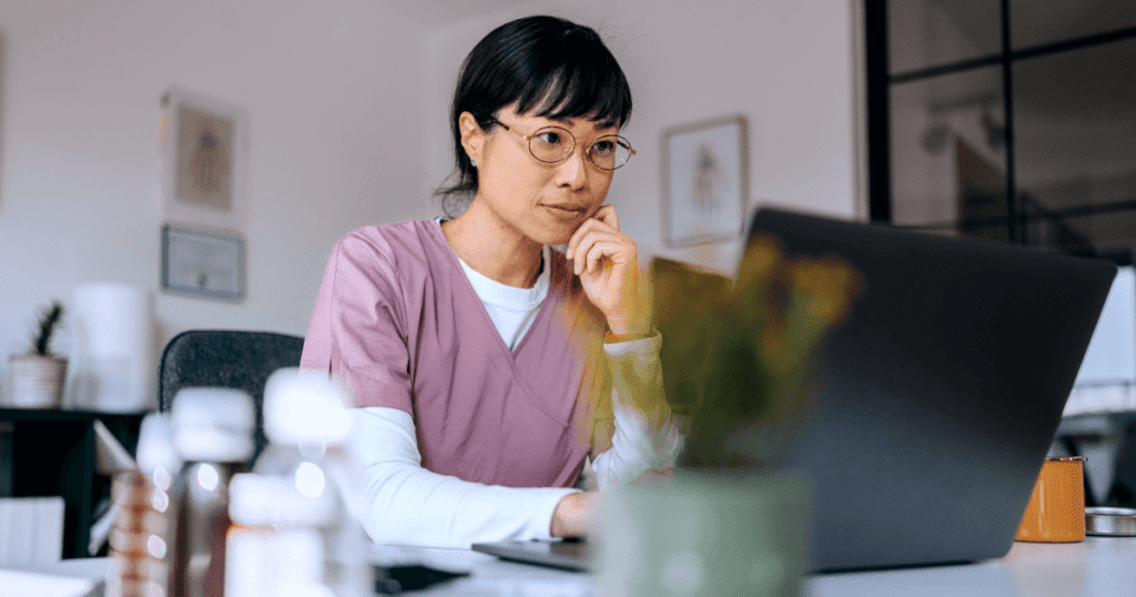 Woman in glasses and pink scrubs thoughtfully reviewing data on a laptop screen