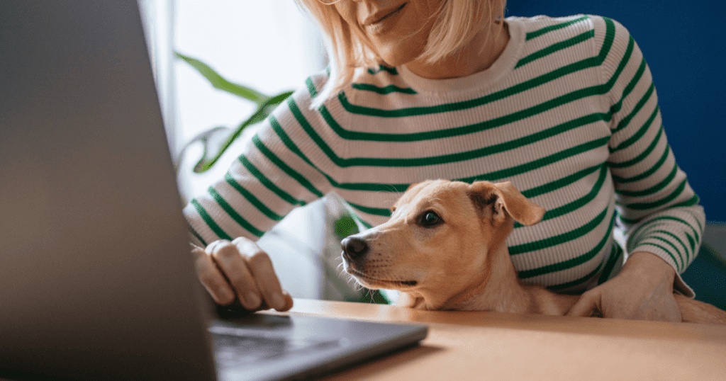 A smiling woman leans over her laptop with her small dog in her lap