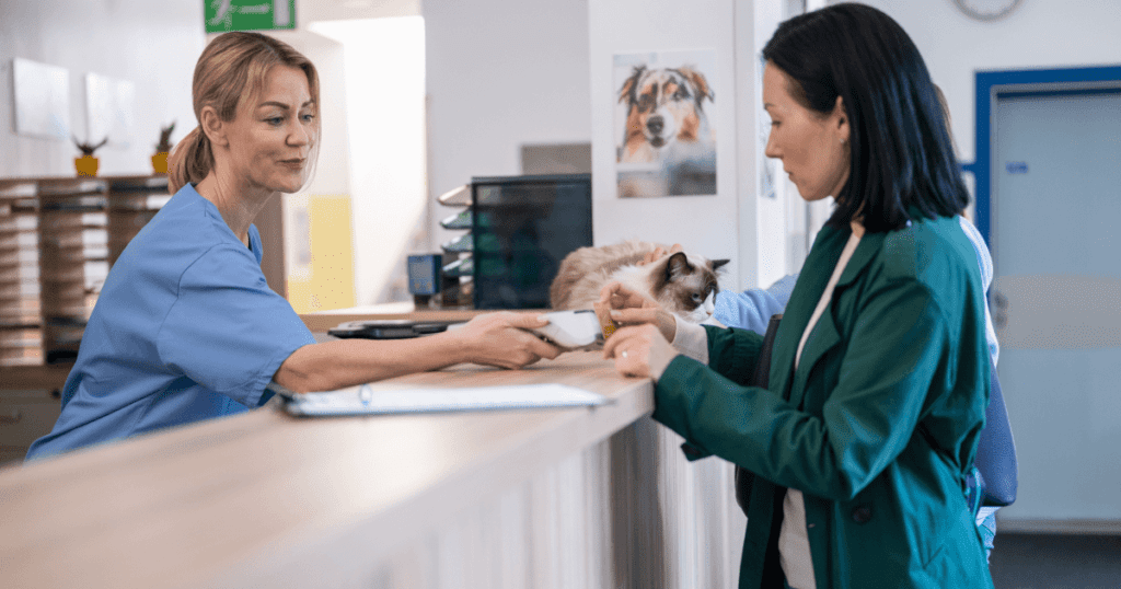 A veterinary practice staffer holds a tap-to-pay reader for a customer to make their payment, with a cat sitting on the counter nearby