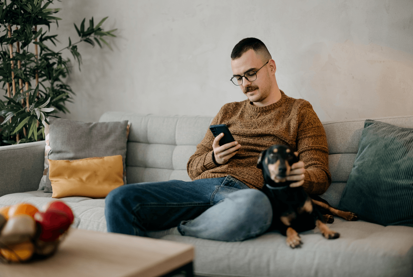 Man on couch viewing his smartphone with small black and brown dog beside him