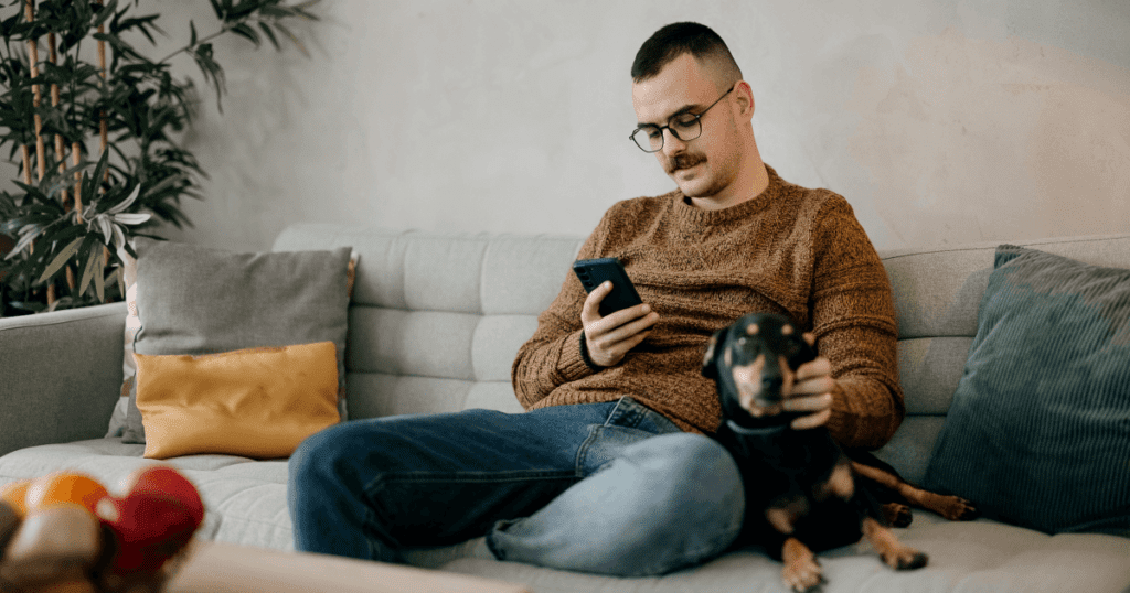 Man on couch viewing his smartphone with small black and brown dog beside him